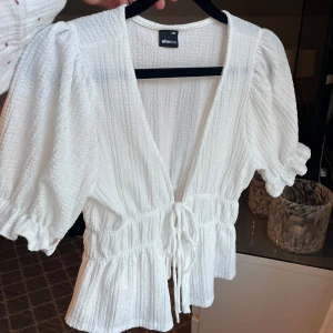 Vit blus med puffärm från Gina Tricot - Supersöt vit topp från Gina Tricot med puffiga korta ärmar, djup v-ringning och knyt i midjan. Toppen har en strukturerad, lite ribbad känsla och volangkant nertill. Perfekt för dig som gillar romantisk och trendig stil. Toppen är använd ett fåtal gången men är fortfarande i superbra skick! 🥰