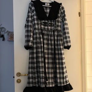 Rutig långklänning från Cheeseday - Supersnygg svartvit rutig långklänning från Cheeseday med stora krage, volangdetaljer och två coola bälten framtill. Klänningen har långa ärmar och markerad midja, samt svart volangkant nertill. Perfekt för dig som gillar statement-plagg med unik stil. 
