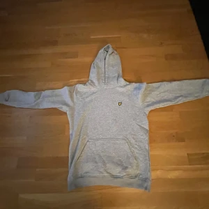 Grå hoodie från Lyle & Scott - Snygg grå hoodie från Lyle & Scott med klassisk gul logga på bröstet. Tröjan har huva, magficka och långa ärmar. Perfekt för chill dagar eller när du vill ha en enkel och stilren look.