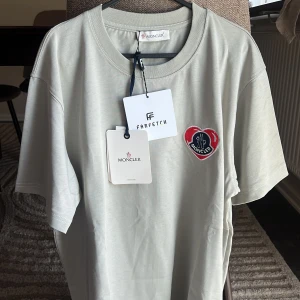 Ljusgrå Moncler t-shirt med hjärtdetalj - Snygg ljusgrå t-shirt från Moncler med klassisk rund hals och korta ärmar. På bröstet sitter en broderad hjärtformad patch i rött och blått med Moncler-logga. Tillverkad i mjuk bomull och har en clean, stilren look som passar till allt.