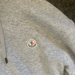 Grå Moncler hoodie med huva - Snygg grå hoodie från Moncler med klassisk logga på bröstet. Tröjan har huva med snörning och ribbade muddar vid ärmslut och nederkant. Perfekt för dig som gillar stilren och bekväm streetwear.