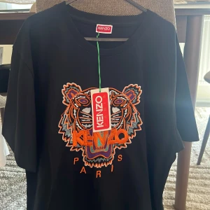 Svart Kenzo t-shirt med tigerbrodyr - Kenzo t-shirt i svart med ikonisk broderad tiger i orange, lila och vitt framtill. Klassisk passform och rund hals. Stora broderade detaljer och logga ger en riktigt fet look. Perfekt för dig som vill sticka ut med streetstyle.