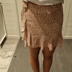 Blommig omlottkjol från SHEIN XS - Supersöt kort kjol från SHEIN i beige med vitt blommönster. Kjolen har omlottmodell med knyt i sidan och volangkant nedtill som ger en extra feminin vibe. Perfekt för varma dagar och lätt att matcha med en enkel topp.