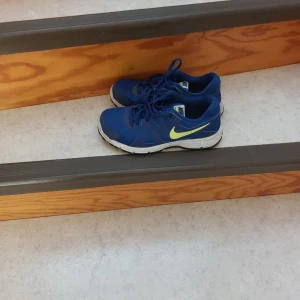 Blå Nike sneakers med neongul swoosh - Snygga blå Nike sneakers med neongul swoosh på sidan och vit sula. Skorna har snörning och en sportig vibe, perfekta för dig som gillar att sticka ut. Ovandelen är i syntetmaterial och sulan är svart undertill för extra grepp.