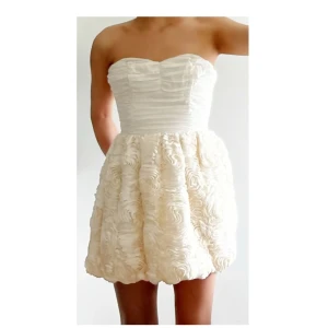 Vit axelbandslös kortklänning - Supersöt axelbandslös kortklänning i vit nyans med voluminös kjol och rosformade detaljer. Överdelen är draperad och figurnära, medan kjolen har en fluffig struktur som ger en romantisk känsla. Perfekt för student eller sommarens fester.