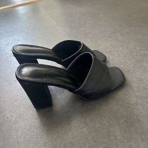 Svarta klackade sandaler med fyrkantig tå - Stilrena svarta sandaler med bred rem över foten och fyrkantig tå. Skorna har hög, kraftig klack och öppen tå, vilket ger en modern och trendig look. Perfekta för dig som vill ha en edgy vibe till outfiten.