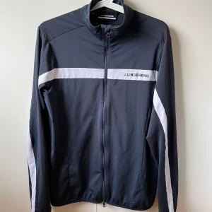 Mörkblå golftröja från J.Lindeberg - Sportig mörkblå tröja från J.Lindeberg med vita ränder över bröstet, armarna och ryggen. Tröjan har hel dragkedja framtill, hög krage och lång ärm. Perfekt för dig som gillar stilren och funktionell design. Väldigt skön och passar lika bra till vardagen som på golfbanan. Storleken är S. Skriv vid eventuella frågor! Pris kan vid snabb affär diskuteras