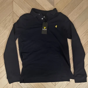 Marinblå pikétröja Lyle & Scott - Snygg marinblå långärmad pikétröja från Lyle & Scott med klassisk krage och två knappar. Tröjan har det ikoniska gula logobroderiet på bröstet och är gjord i skönt material. Perfekt för en stilren och avslappnad look. Vi frågor eller fler bilder kan ni höra av er. Passar mig som är 173 perfekt så passar nog både S och XS!