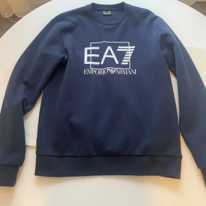 Mörkblå EA7 Emporio Armani sweatshirt - Snygg mörkblå sweatshirt från EA7 Emporio Armani med stort vitt logotryck på bröstet. Tröjan har rund hals, långa ärmar och ribbade muddar. Perfekt för dig som gillar stilrena och exklusiva plagg med streetkänsla.