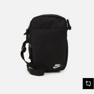 Svart axelväska från Nike - Snygg svart axelväska från Nike med justerbar rem och två praktiska fack med dragkedja. Väskan har en stilren design med vit Nike-logga framtill och är tillverkad i slitstark polyester. Perfekt för att bära med sig det viktigaste på stan eller till träningen.