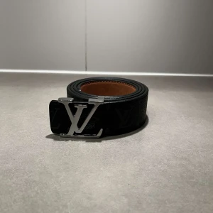 Svart Louis Vuitton skärp med LV-spänne - Säljer ett svart Louis Vuitton bälte i skinn med klassiskt LV-monogram och silverfärgat LV-spänne. Bältet har en bred design och insidan är brun. Perfekt accessoar för att lyfta din outfit med en lyxig touch.