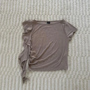 Beige topp med volang från Gina Tricot - Säljer en beige topp från Gina Tricot i storlek L (passar mig som har M) Toppen har kort ärm på ena sidan och en cool volangdetalj med snörning längs andra sidan. Mjuk och stretchig känsla, perfekt för dig som gillar unika detaljer. Nästan aldrig använd
