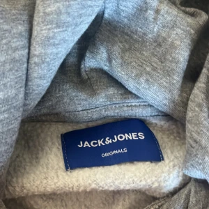 Grå hoodie från Jack & Jones i storlek S - Snygg grå hoodie från Jack & Jones med tryck framtill i blått och vitt. Tröjan har huva med snörning och är tillverkad i mjuk bomullsblandning med fleecefoder på insidan. Perfekt för chill dagar och streetstyle. Storlek S