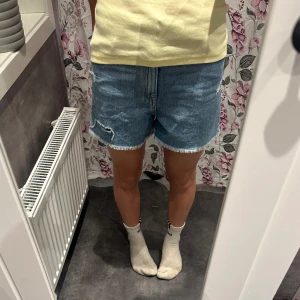 Blå jeansshorts med fransig kant - Snygga blå jeansshorts med fransig kant och slitna detaljer. Klassisk femficksmodell med bälteshällor och justerbar midja på insidan. Perfekta för en avslappnad stil under varma dagar. Jag kommer inte riktigt ihåg men tror shortsen är från H&M