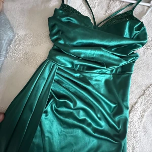 Grön långklänning med snörning - Snygg grön långklänning i glansigt satinliknande material med smala axelband och v-ringad urringning. Klänningen har draperade detaljer framtill och snörning i ryggen för en justerbar passform. Perfekt för dig som vill sticka ut med en elegant look.