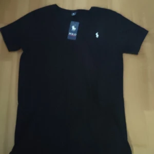 Svart t-shirt från Polo Ralph Lauren - Svart t-shirt från Polo Ralph Lauren med klassisk vit broderad logga på bröstet. T-shirten har rund halsringning och korta ärmar. Tillverkad i mjuk bomull för en skön känsla och clean look. Perfekt för dig som gillar stilrena basics med premiumkänsla.