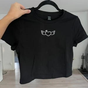 Svart croppad topp med strasshjärta - Svart croppad topp från Shein med korta ärmar och ett glittrigt strassmotiv av ett hjärta med vingar på bröstet. Tillverkad i mjuk bomull med lite stretch för en bekväm passform. Liten i storlek!!! Passar S/M!!! Önskar du veta exakta mått? Tveka inte att höra av dig!