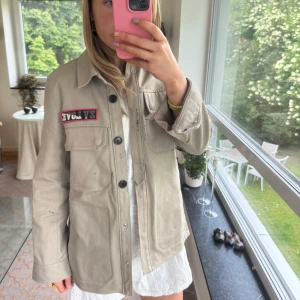 Overshirt från Zadig & Voltaire med patches - Beige overshirt från Zadig & Voltaire med coola patches och tryck. Framsidan har en ZV LOVE-patch och ärmen har en döskalle med flames. Baksidan har camomönster och guldstjärnor. Jackan har knappar framtill och stora fickor. Perfekt för en edgy streetstil.