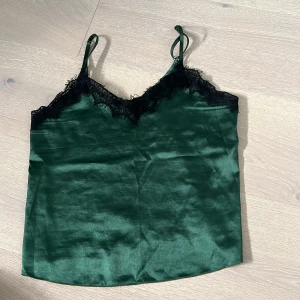 Grön satinlinnetopp med spets - Mörkgrön linnetopp i satin med tunna axelband och svart spets längs v-ringningen. Snygg glansig finish och feminin känsla, perfekt att styla med jeans eller kjol. Lätt och luftig, passar dig som gillar trendiga detaljer.