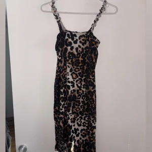Leopardmönstrad kort klänning - Snygg kort klänning med leopardmönster och smala axelband med twistad detalj. Klänningen har en rak passform och är i mjukt, lätt material. Färgerna går i svart, beige och brunt, vilket ger en cool och trendig look.