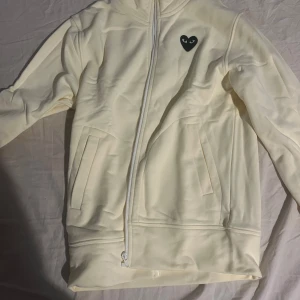 Vit hoodie från Comme des Garçons Play - Snygg vit hoodie från Comme des Garçons Play med dragkedja och huva. Ikoniskt svart hjärta med ögon på bröstet och stort hjärta på ryggen. Mjuk och skön i bomull, perfekt för en clean och stilren look.