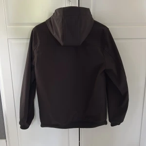 Svart vindjacka med luva - Snygg svart vindjacka med luva och långa ärmar. Jackan har en stilren design utan synliga detaljer på ryggen och är tillverkad i ett slätt syntetmaterial. Perfekt för dig som gillar en clean och väldigt snygg look. Jackan är i stort sätt oanvänd och är i nyskick med både fickor med dragkedja och inne fickor.Jackan är köpt på kidsbrand store och har storlek 15-16 år.