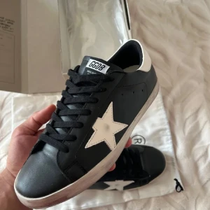 Golden Goose svarta sneakers med stjärna - Golden Goose sneakers i svart skinn med vit stjärndetalj på sidan och vit hälkappa. Klassisk låg modell med svarta skosnören och slitna detaljer på sulan för en cool, vintage look. Perfekta för dig som gillar streetstyle och vill sticka ut.Bästa pris någonsin