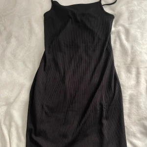 Svart ribbad kortklänning med öppen rygg - Snygg svart ribbad kortklänning med smala axelband och öppen rygg med knyt. Klänningen har en tight passform och är tillverkad i ett stretchigt material som sitter skönt på kroppen. Perfekt för dig som gillar minimalistisk och stilren look.