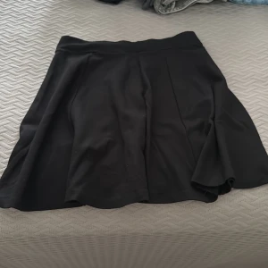 Svart kortkjol med dragkedja - Snygg svart kortkjol med markerad midja och silvrig dragkedja bak. Kjolen har en lätt utsvängd form som ger en skön vibe och passar perfekt till sneakers eller boots. Enkel och stilren design utan mönster eller extra detaljer.