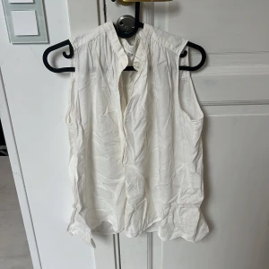 Vit ärmlös blus från Uniqlo - Säljer en luftig och stilren vit blus från Uniqlo i storlek M. Blusen är ärmlös med lätt rynkade axlar och har en klassisk krage med knäppning framtill. Perfekt för en fräsch och enkel look under varmare dagar.