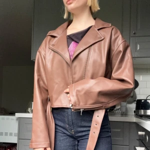 Brun skinnjacka från Nelly x Bianca - Snygg brun skinnjacka från Nelly med croppad passform, bred krage och detaljer som bälte i midjan och dragkedja framtill. Jackan har långa ärmar och markerade sömmar som ger en trendig look. En något större passform.