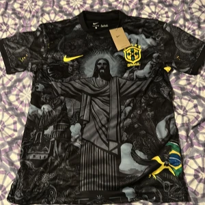 Nike Brasil svart fotbollströja - Unik svart Brasilien fotbollströja från Nike med stort motiv av Kristusstatyn och detaljerade grafiska inslag. Tröjan har korta ärmar, broderat CBF-märke och gula Nike-loggor. Materialet är lätt och andas, perfekt för match eller streetwear.