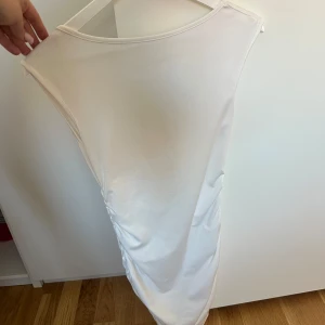 Vit ärmlös långklänning med rynk - Snygg vit långklänning med djup urringning i ryggen och rynkade detaljer längst sidorna. Klänningen är ärmlös och har en tight passform som framhäver figuren. Perfekt för dig som vill sticka ut med en stilren look.