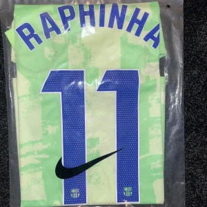 Barcelona Raphinha bortatröja Nike M - Barcelona bortatröja med Raphinha 11-tryck. Tröjan är ljusgrön med abstrakt mönster, blå detaljer och klubbmärke på bröstet. Nike-logga och Spotify-sponsor framtill. Tillverkad i lätt och ventilerande material, perfekt för match eller träning.