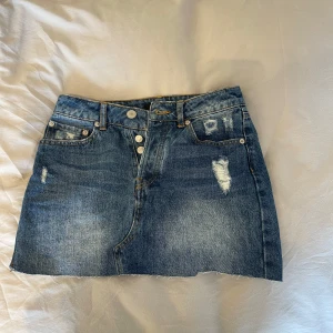 Jeanskjol från BikBok med slitningar - Snygg blå jeanskjol från BikBok i kort modell med rå kant nertill och slitna detaljer framtill. Kjolen har klassiska fickor, knappar och en cool, avslappnad vibe. Perfekt för dig som gillar streetstyle och denim.