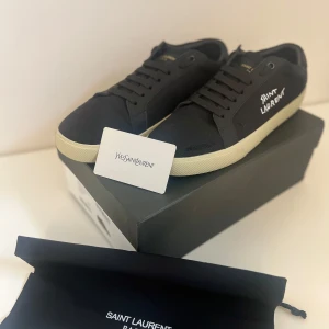 Svarta Saint Laurent sneakers strl 42 - Snygga svarta sneakers från Saint Laurent med vit logga broderad på sidan och gulddetaljer på plösen. Skorna har en klassisk låg profil, svarta snören och vit platt sula. Tillverkade i canvas och skinn för en stilren och lyxig känsla.