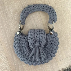 Handgjord grå stickad handväska - Unik handväska i grått, virkad i tjockt garn med rund form och dekorativ tofs framtill. Väskan har ett kort handtag och stängs med magnetlås. Snygga silvriga metalldetaljer ger extra edge. Perfekt för dig som vill ha något annorlunda och personligt.