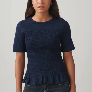 Mörkblå topp med volangkant - Supersnygg mörkblå topp med smockad midja och volangkant nertill. Modellen har rund halsringning och korta ärmar. Perfekt för dig som gillar en feminin och trendig stil.