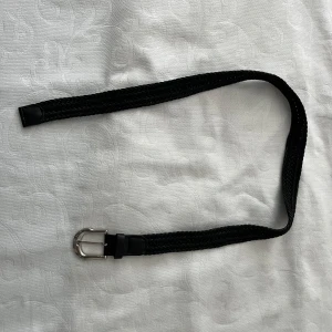 Svart flätat bälte - Oanvänt snyggt svart flätat bälte med ett stilrent silverfärgat spänne. Bältet har detaljer i svart skinn vid ändarna och passar perfekt till jeans eller kostymbyxor. Enkel och tidlös design som lyfter vilken outfit som helst. Ca 100 cm.