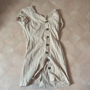 Beige kortklänning med knappar - Snygg beige kortklänning med korta ärmar och knappar längs hela framsidan. Klänningen har en enkel, rak passform och är tillverkad i ett lätt linneliknande material. Perfekt för varma dagar och enkel att styla med accessoarer.
