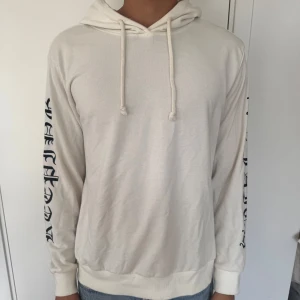 Vit hoodie med tryck på ärmarna - Säljer en vit hoodie med huva och snörning. Tröjan har svart tryck i gotisk stil längs båda ärmarna. Mjuk och skön, perfekt för en chill look. Passar dig som gillar streetwear och enkel stil.