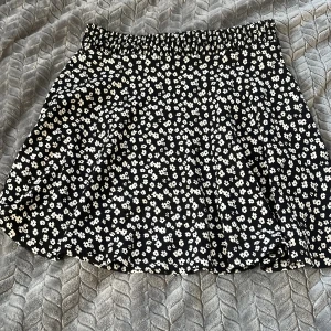 Svart blommig kortkjol från 157 - Supersöt svart kortkjol från 157 med vita småblommor över hela tyget. Kjolen har resår i midjan för en bekväm passform och är perfekt för dig som gillar en feminin och lekfull stil.