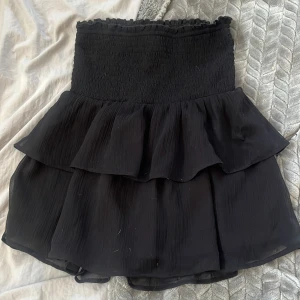 Svart volangkjol från Gina Tricot - Supersöt svart kortkjol från Gina Tricot med smockad midja och dubbla volanger. Kjolen är luftig och har en lätt, flowy känsla som gör den perfekt för en trendig look. Passar dig som gillar att sticka ut med stilrena detaljer. Aldrig använd. Ordenarie pris 300