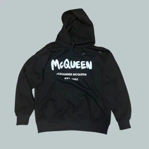 Svart hoodie från Alexander McQueen - Svart hoodie från Alexander McQueen med stor vit logga och text på bröstet. Klassisk huva med snörning och känguruficka framtill. Perfekt för dig som gillar streetwear och vill sticka ut med en ikonisk design.