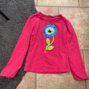 Rosa långärmad tröja med paljettblomma str 134 / 140 - Snygg rosa långärmad tröja från Kids by Kappahl med en stor, färgglad blomma i paljetter på bröstet. Blomman har blå, gröna och rosa detaljer som verkligen poppar. Perfekt för dig som gillar att sticka ut med coola detaljer.