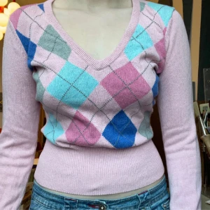 Rutig rosa v-ringad stickad tröja - Supersöt stickad tröja i rosa med rutigt mönster i blått, grått, ljusblått och cerise framtill. V-ringad och figurnära passform med långa ärmar och bred ribbad mudd i midjan. Perfekt för dig som gillar preppy vibes och färg!