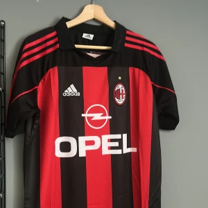 AC Milan Inzaghi 9 Adidas fotbollströja - Klassisk AC Milan fotbollströja med Inzaghi 9 på ryggen. Tröjan är svart och röd med breda vertikala ränder, Adidas-logga och Opel som sponsor framtill. Korta ärmar och klubbmärke på bröstet. Perfekt för dig som älskar retro och italiensk fotboll.helt ny med tagg en replika