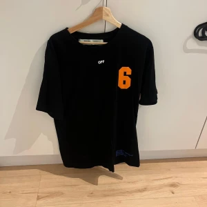 Svart Off-White t-shirt med orange tryck - Svart t-shirt från Off-White med stort orange tryck på ryggen och en orange siffra 6 på bröstet. Klassisk rund hals och korta ärmar. Materialet är mjuk bomull och passformen är normal. Snygg streetwear-vibe med tydlig logga och grafiskt motiv.