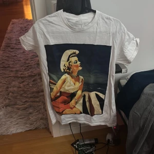 Vit t-shirt från Revenite - Säljer en vit t-shirt med ett coolt retrotryck av en kvinna i solhatt. Perfekt för en avslappnad stil. T-shirten är kortärmad och gjord i mjukt material. Varan är från det eleganta merket Revenite