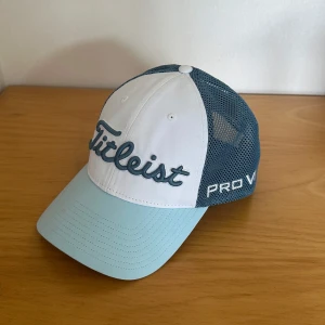 Titleist golf keps  - Snygg golf keps från Titleist med vit front, ljusblå skärm och blå mesh på sidor och bak. Broderad Titleist-logga framtill och Pro V1-text på sidan. Perfekt för golfbanan eller en sportig look. Justerbar passform och ventilerande material.